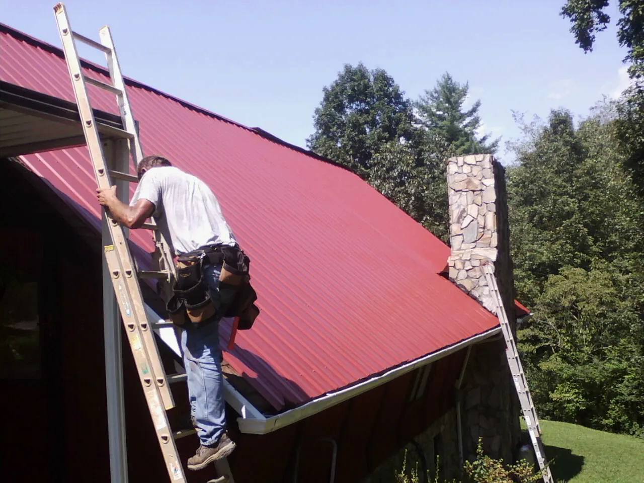 Roofer on a ladder beside a red metal roof and stone chimney — Soffit & Fascia Repair by Roofing Brothers in Shelley